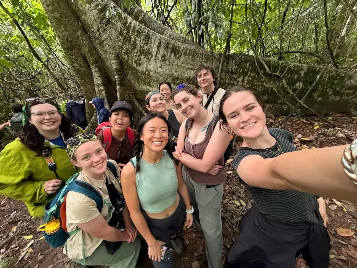 A group selfie of smiling young adults by the massive buttress roots of a Chilamate tree in a Costa Rican rainforest.