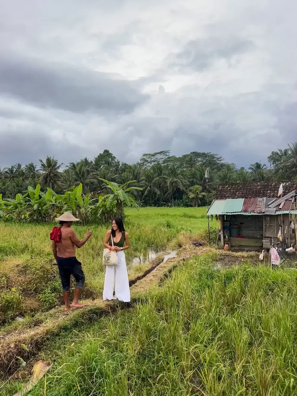 Eugenia talks happily with a local farmer who is giving her a thumbs up in a green rice paddy field.