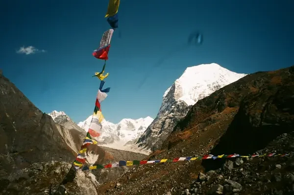 Colorful Buddhist prayer flags string across a rocky hillside, framing jagged, snow-dusted mountain peaks against a deep blue sky.