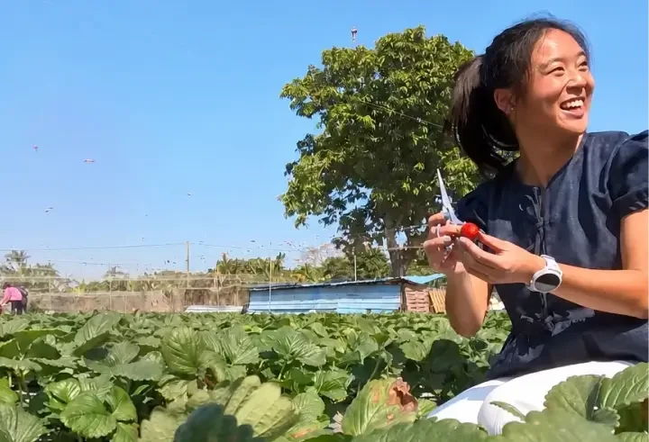 Eugenia laughing while holding pruning shears and a freshly harvested red strawberry in a green farm field under a bright blue sky.