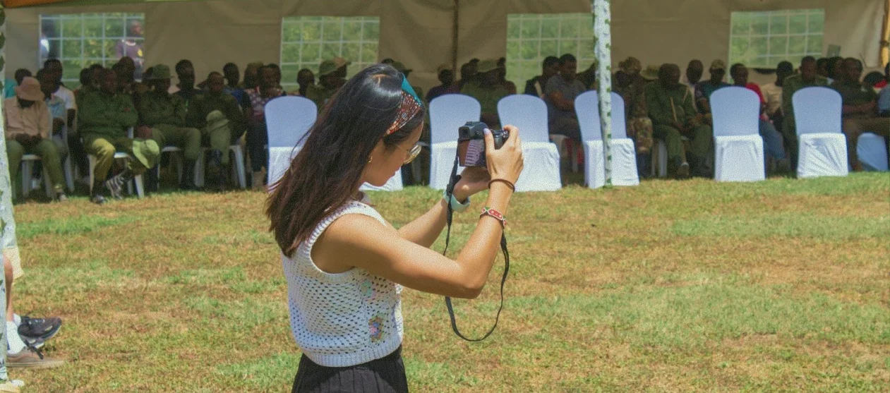 Eugenia holding up a camera to photograph an outdoor community gathering, illustrating environmental media and fieldwork in action.