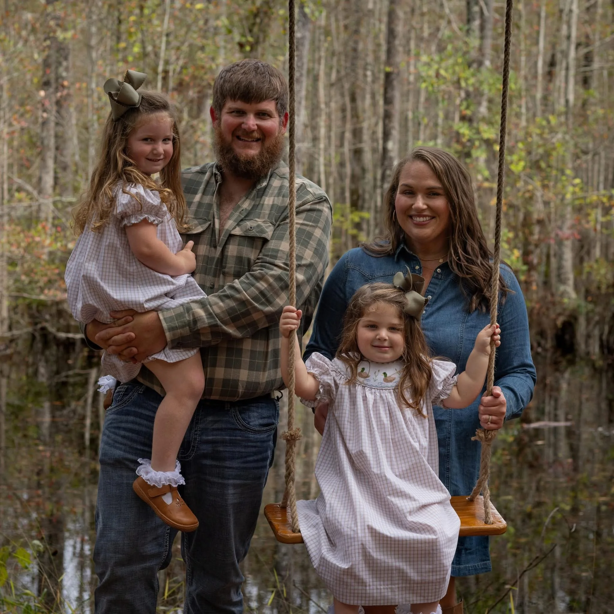 A person in a plaid shirt is standing next to a swing and holding a young girl in a checkered dress on their knee, with two other women nearby, all smiling in a wooded outdoor setting.