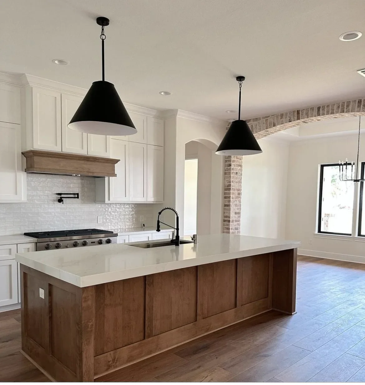 Modern kitchen with white cabinetry, a large island with a wooden base and white countertop, black pendant lights, and a white tiled backsplash. There is a black faucet and a built-in stove, with a brick archway and window in the background.