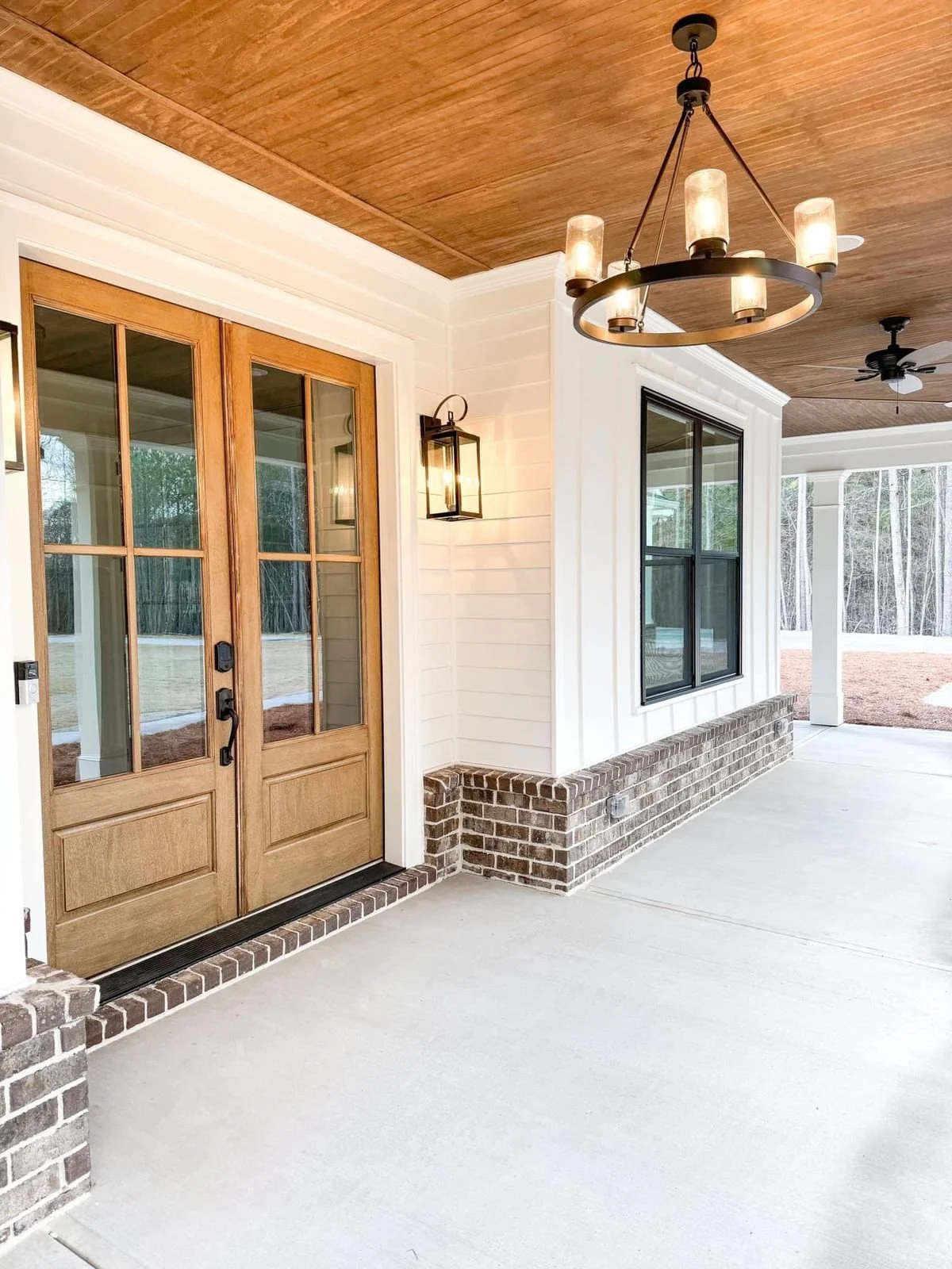 Front porch of a house with wooden double doors, white siding, black-framed window, brick accents, and a ceiling fan with a chandelier.