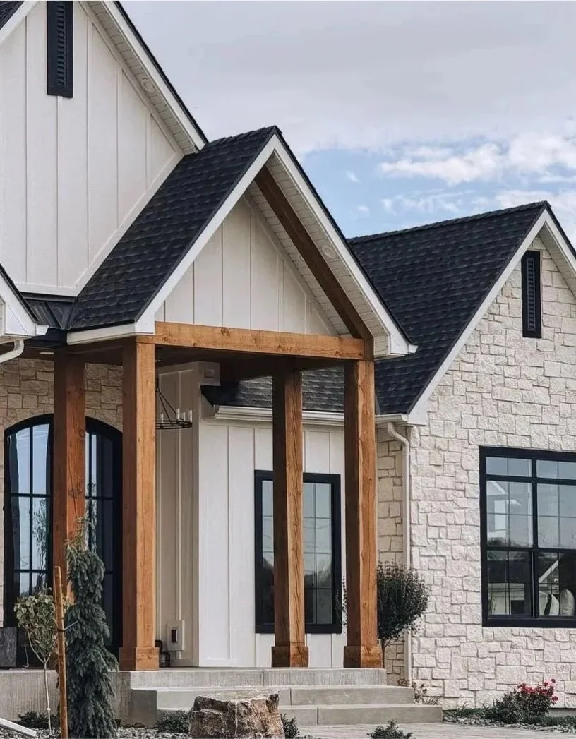 Front view of a house under construction with a wooden porch frame, black shingle roof, white exterior walls, and large black-framed windows, with steps and landscaping in the foreground.