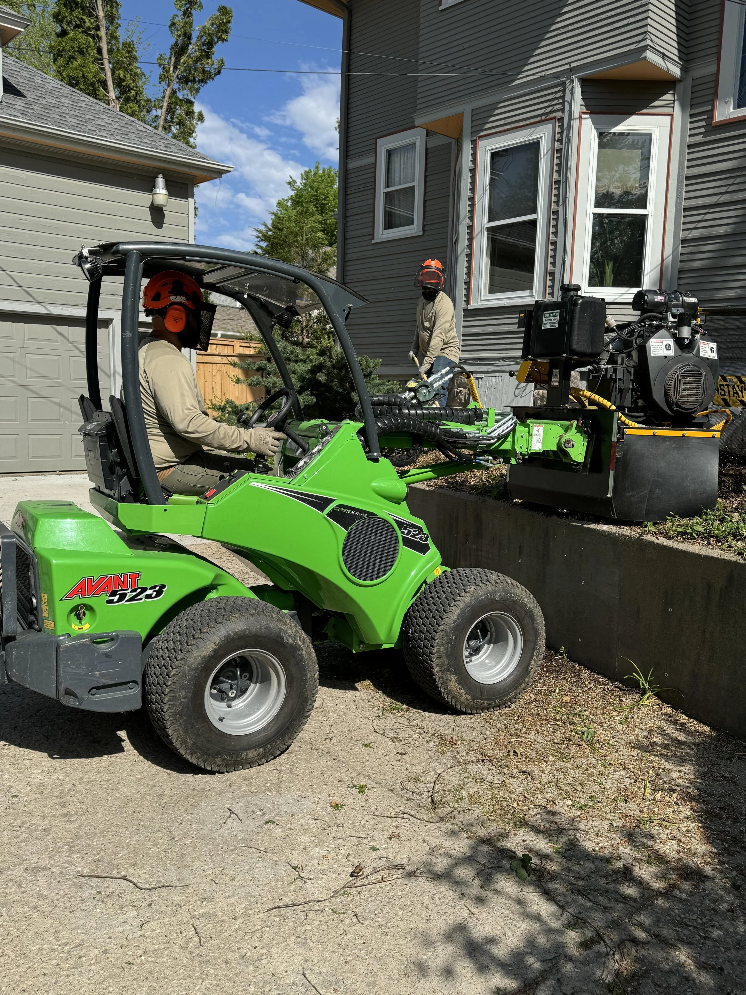 Two workers operating heavy machinery near a house, with one inside a green skid-steer loader and the other on a raised platform, both wearing safety helmets and gloves.