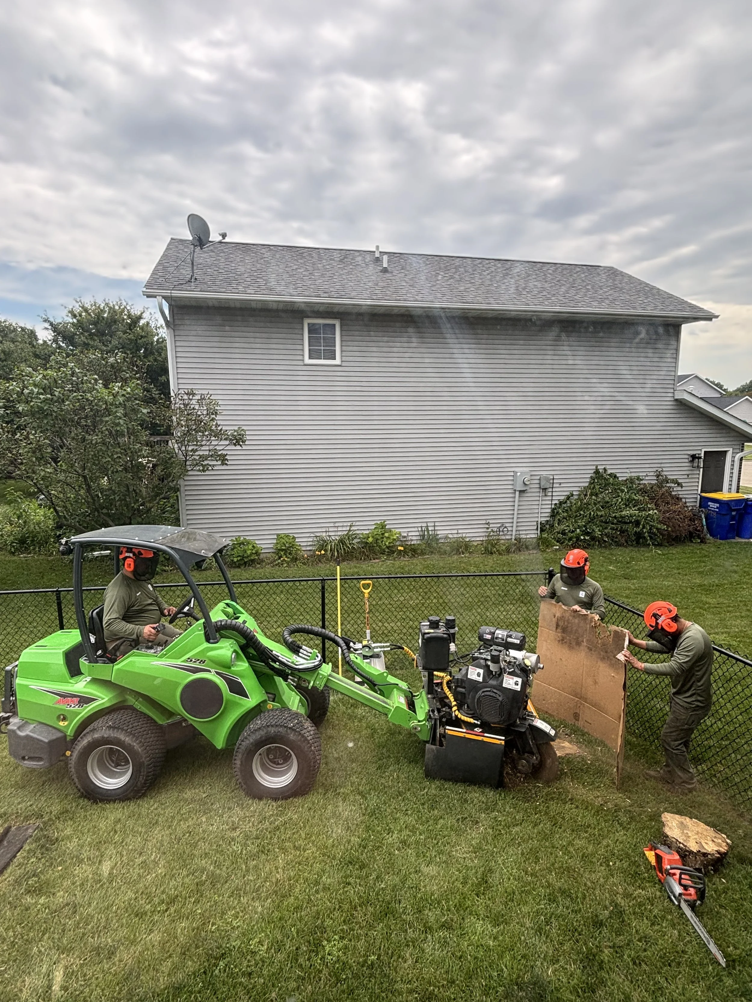 Workers in safety helmets operating a green compact utility tractor with a hydraulic wood splitter attachment on a residential lawn, with a gray house and cloudy sky in the background.