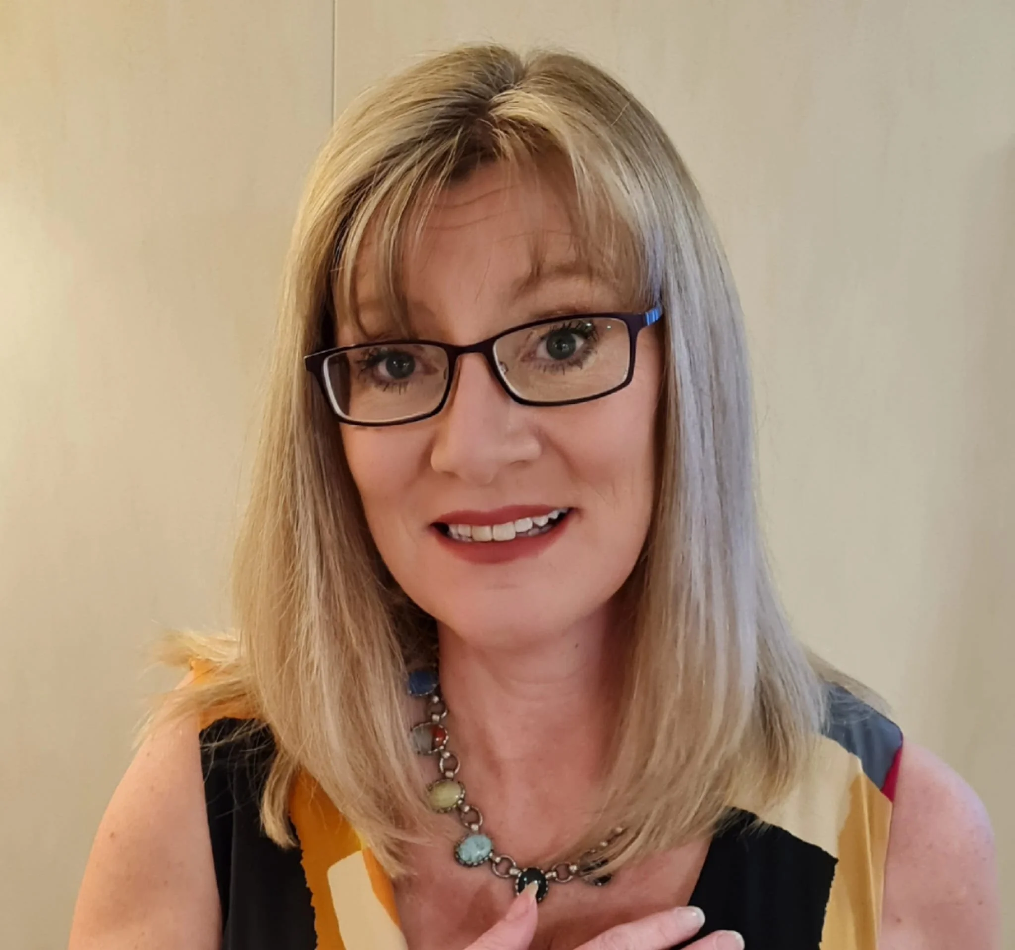 Close-up of a woman with blonde hair, glasses, and a colorful necklace, smiling and touching her chest.