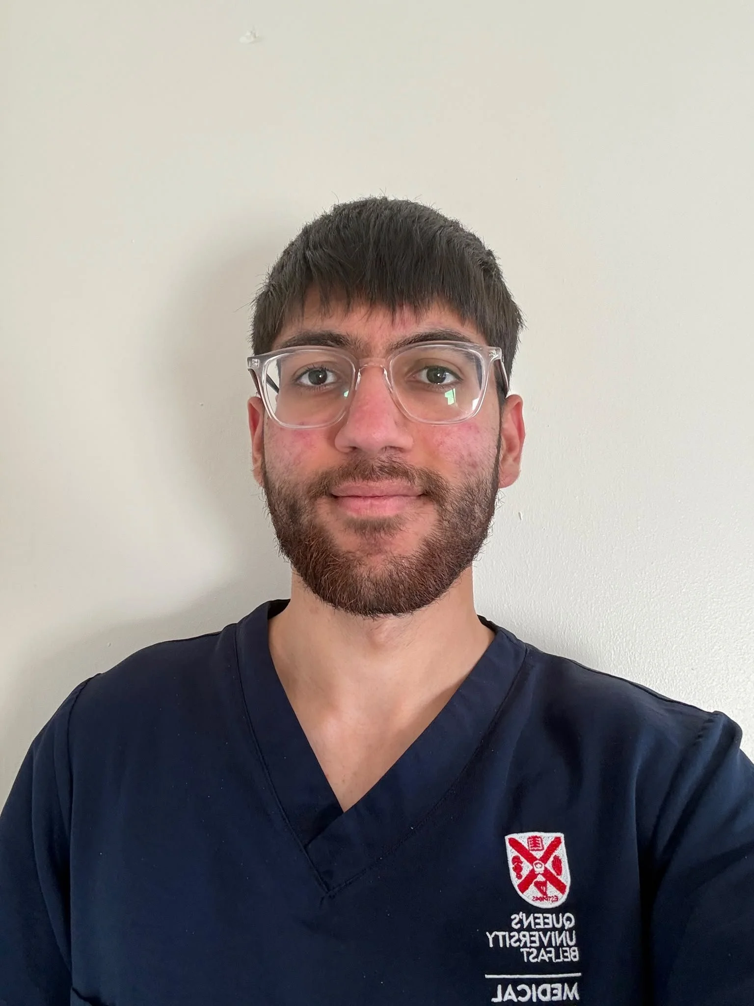 A young man with short dark hair and glasses took a selfie against a plain white wall background. He is wearing navy scrubs with a logo that says Queen's University Unversity and St. Mary's on the chest.