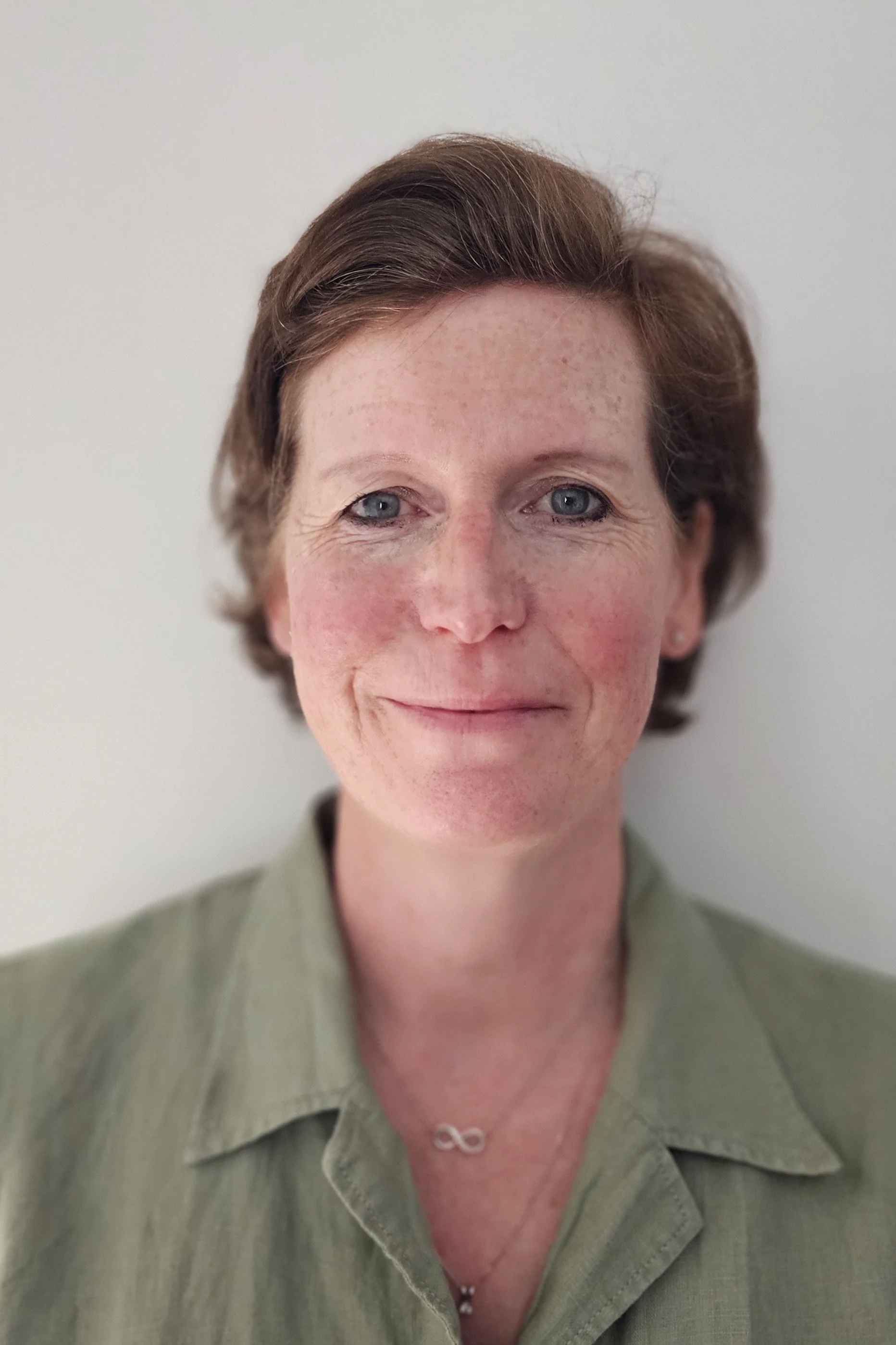 A woman with short brown hair, blue eyes, and light skin, smiling slightly, wearing a green shirt with an infinity symbol necklace, against a plain white background.
