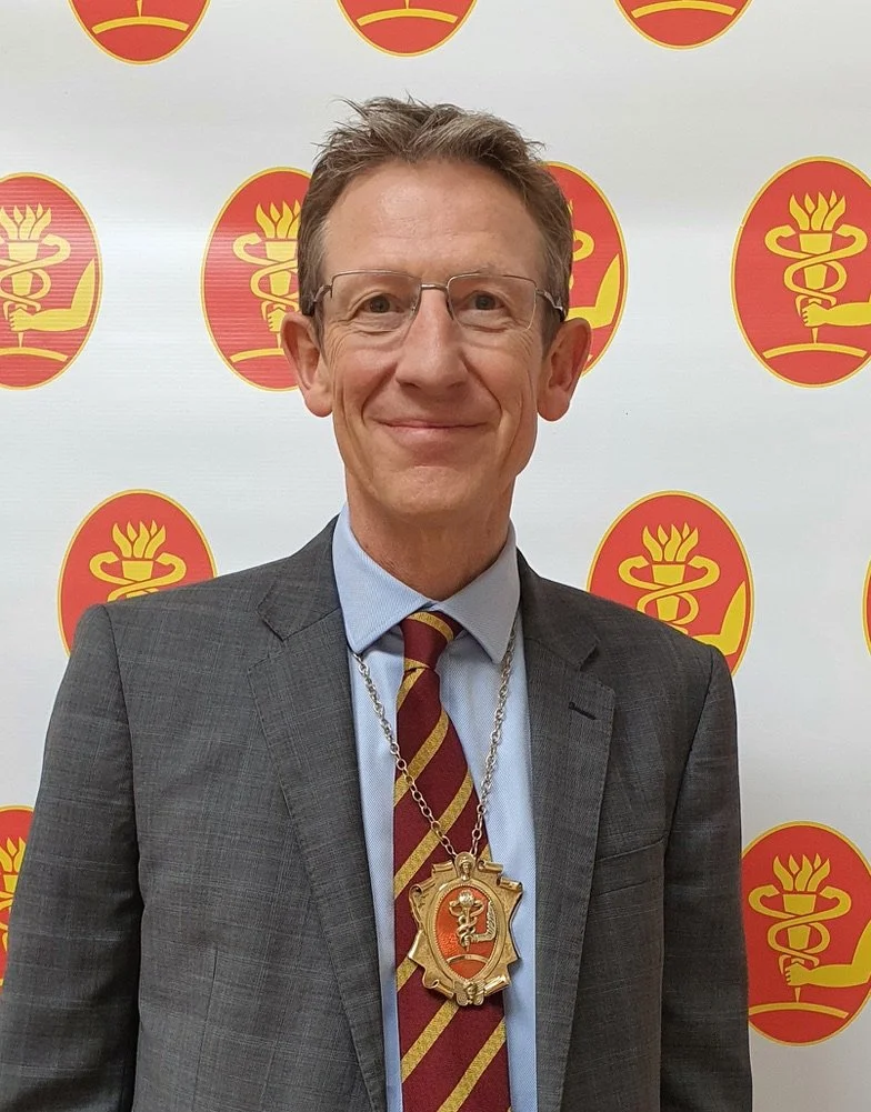 Man in suit with a medal around his neck, standing in front of a backdrop with a repeating logo featuring a stylized snake wrapped around a staff with a flame on top.