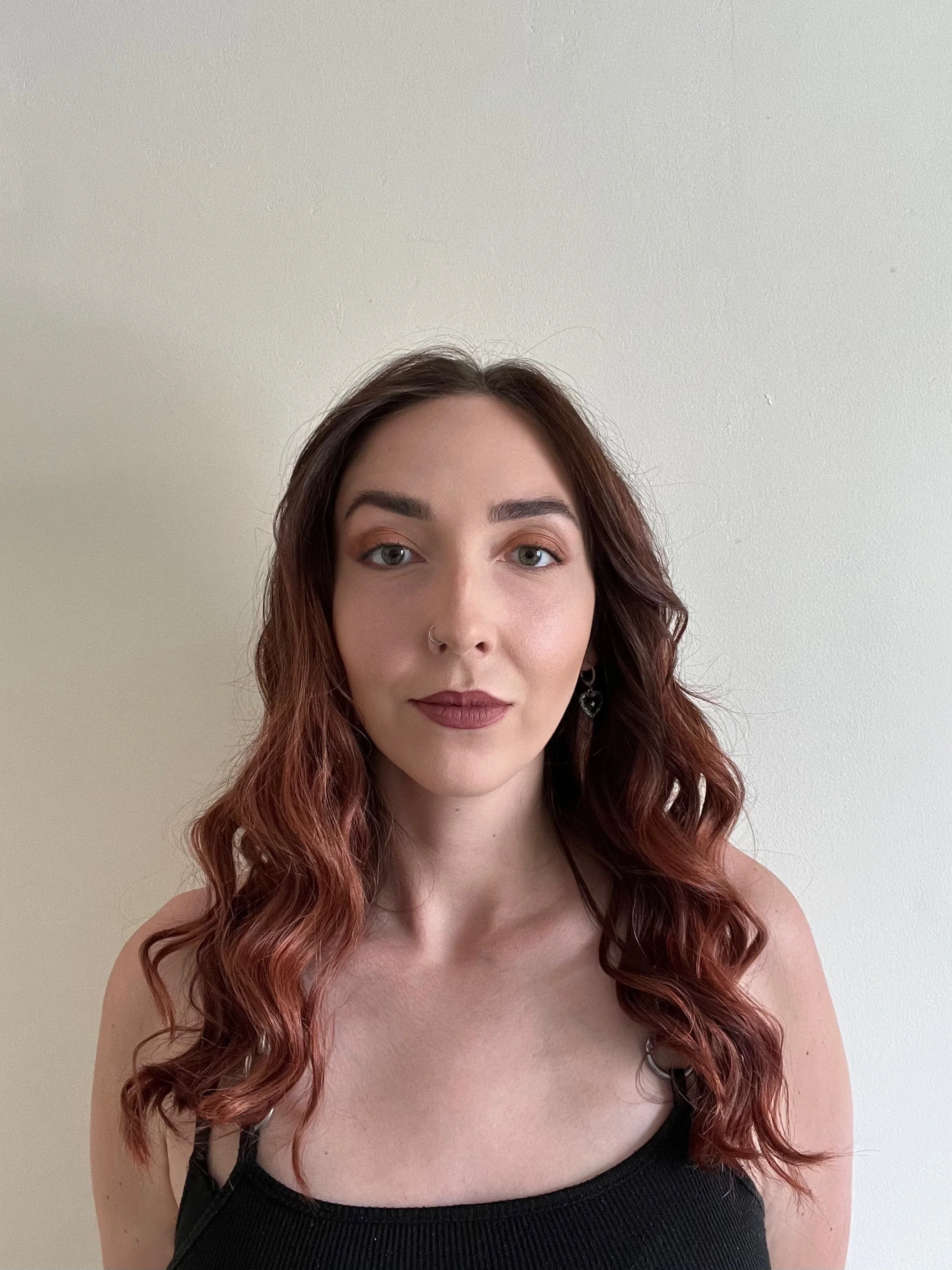 A young woman with long, wavy, reddish-brown hair wearing a black tank top, standing against a plain white wall.
