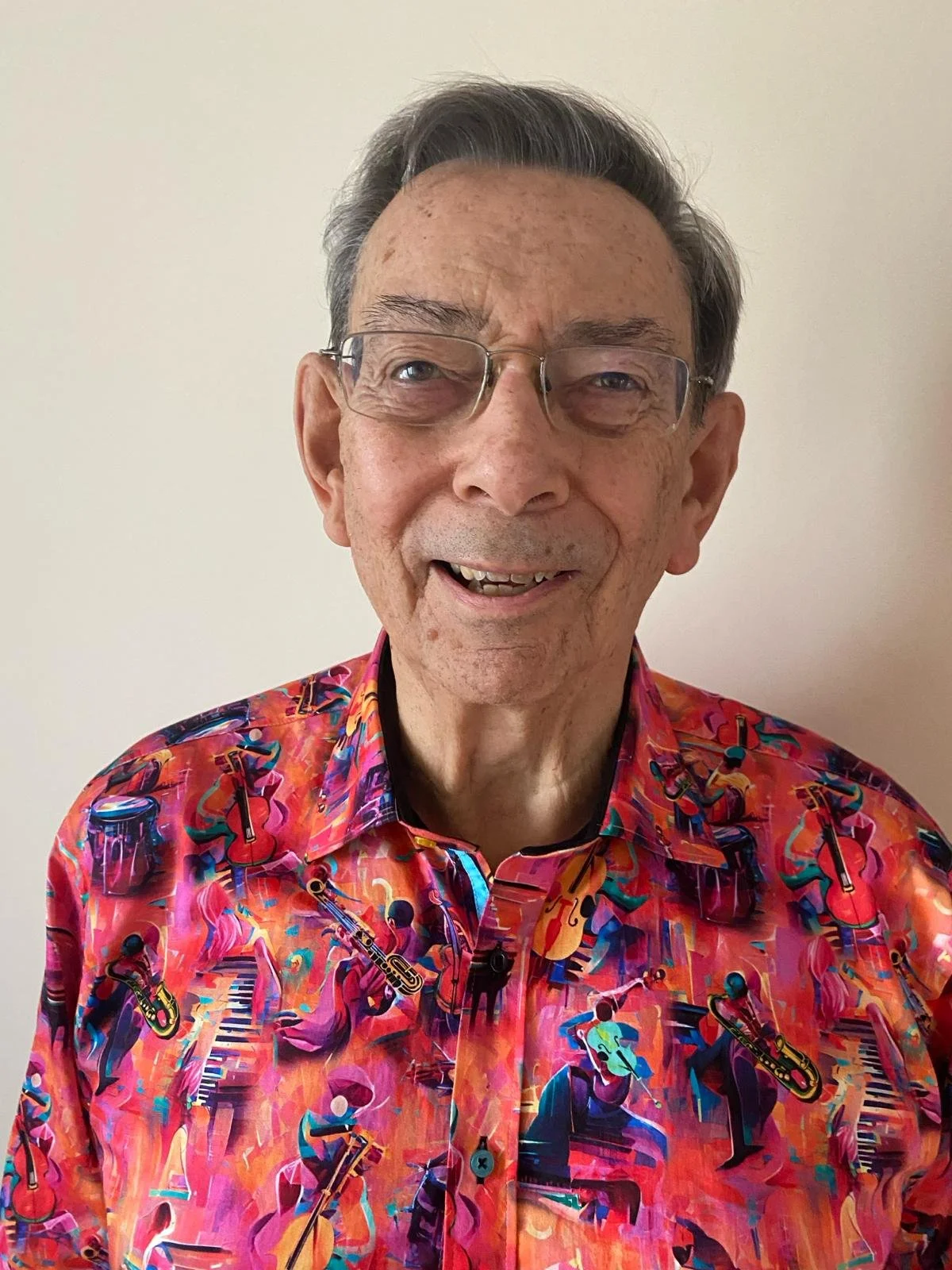 An elderly man with glasses smiling, wearing a colorful shirt with musical instrument patterns, against a plain light-colored background.