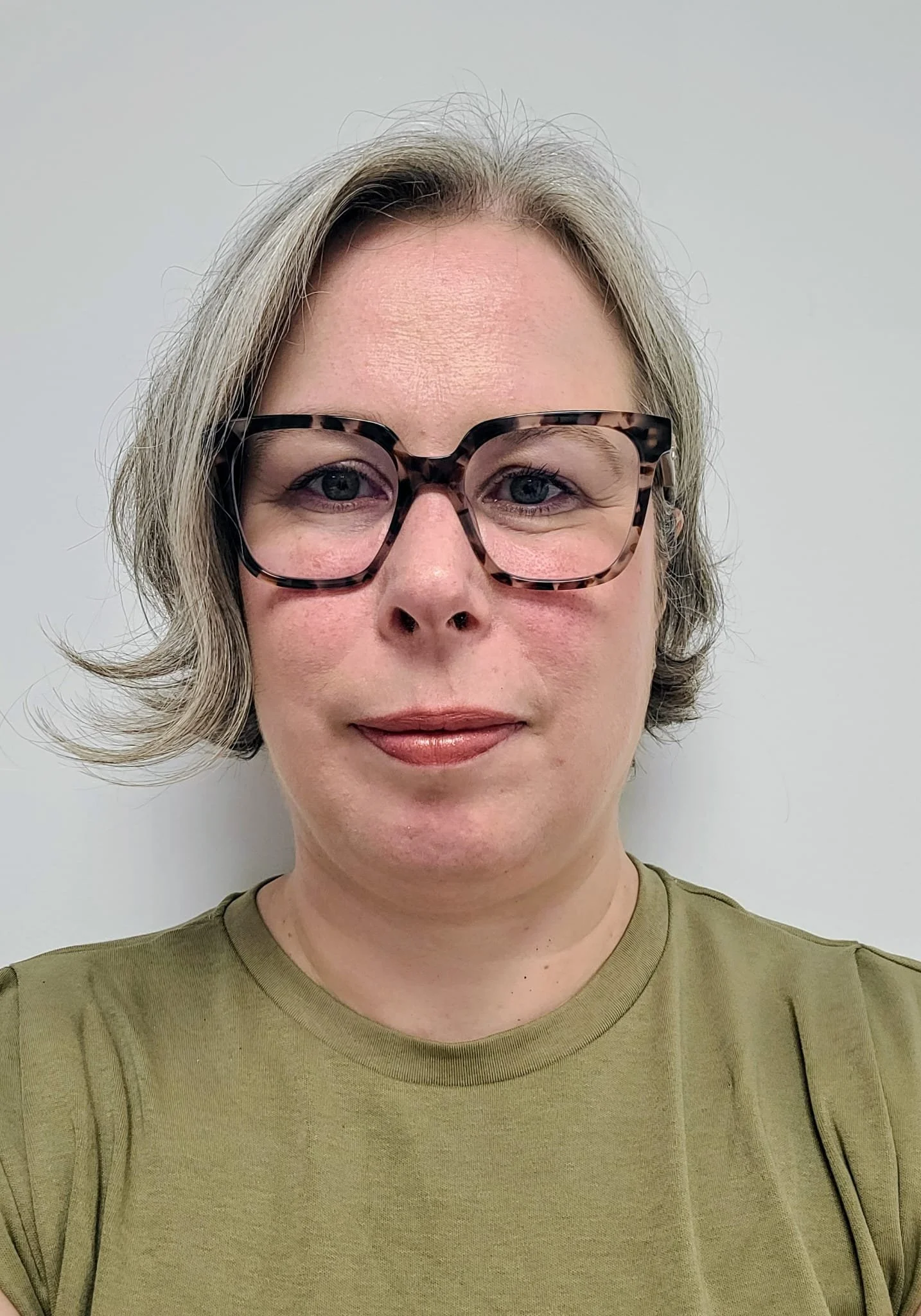 A woman with short gray hair wearing tortoiseshell glasses and an olive green t-shirt standing against a plain light gray background.