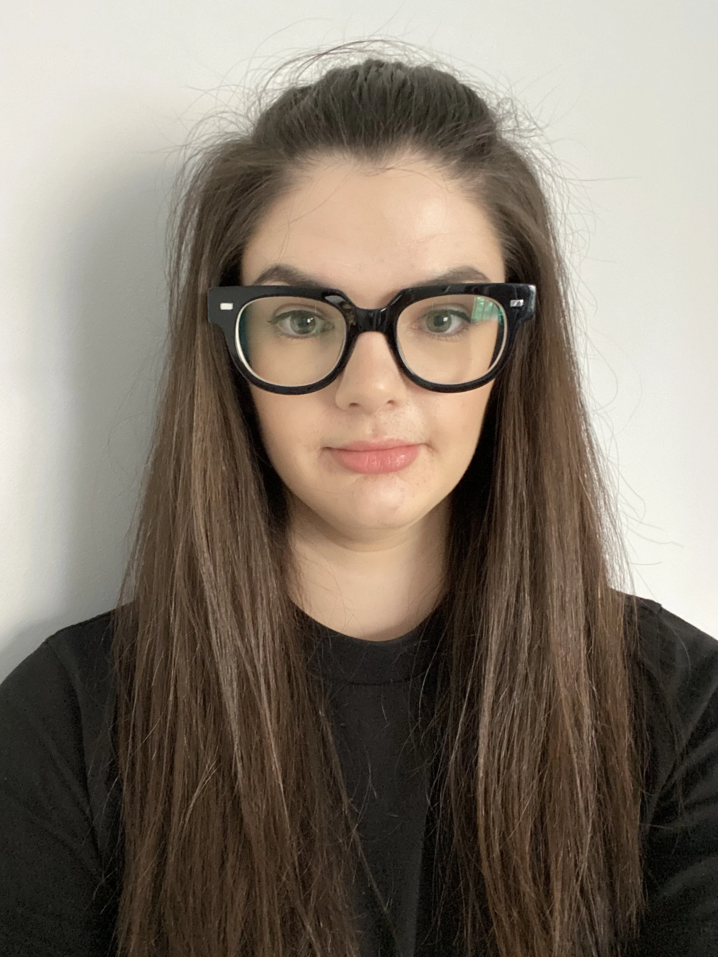A woman with long brown hair wearing black-rimmed glasses and a black top, standing against a plain light-colored wall.