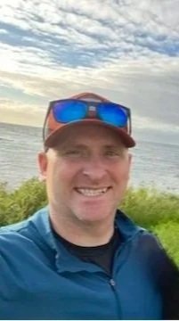 A man outdoors near a body of water on a cloudy day, wearing a red cap, sunglasses on his head, and a blue jacket, smiling at the camera.