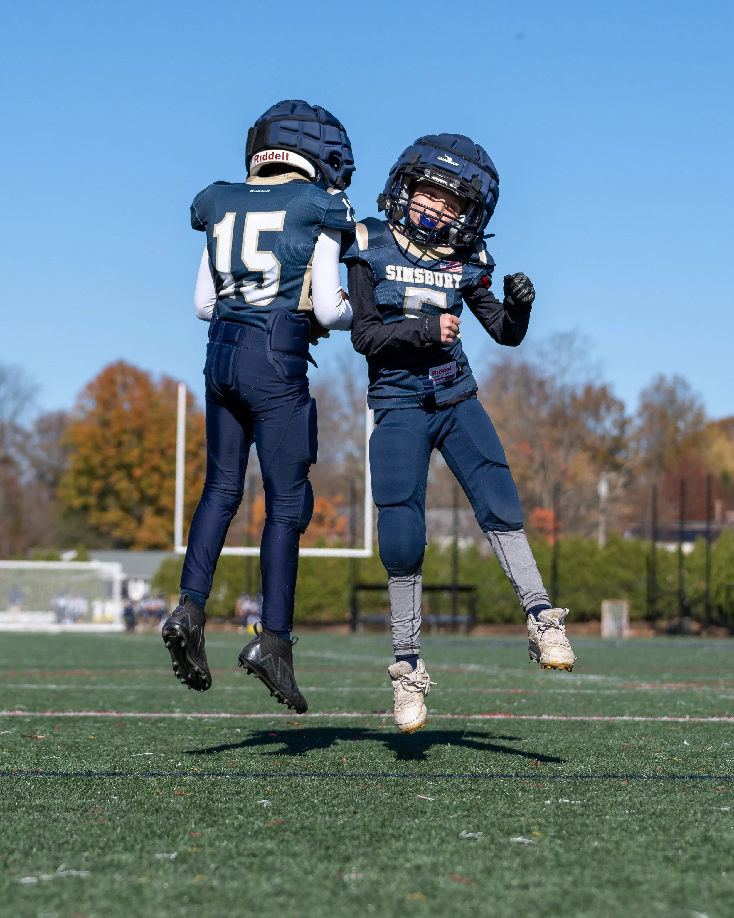Two young football players in a celebratory stance on a field, wearing navy blue and beige uniforms with helmets, with a backdrop of trees and a clear blue sky.