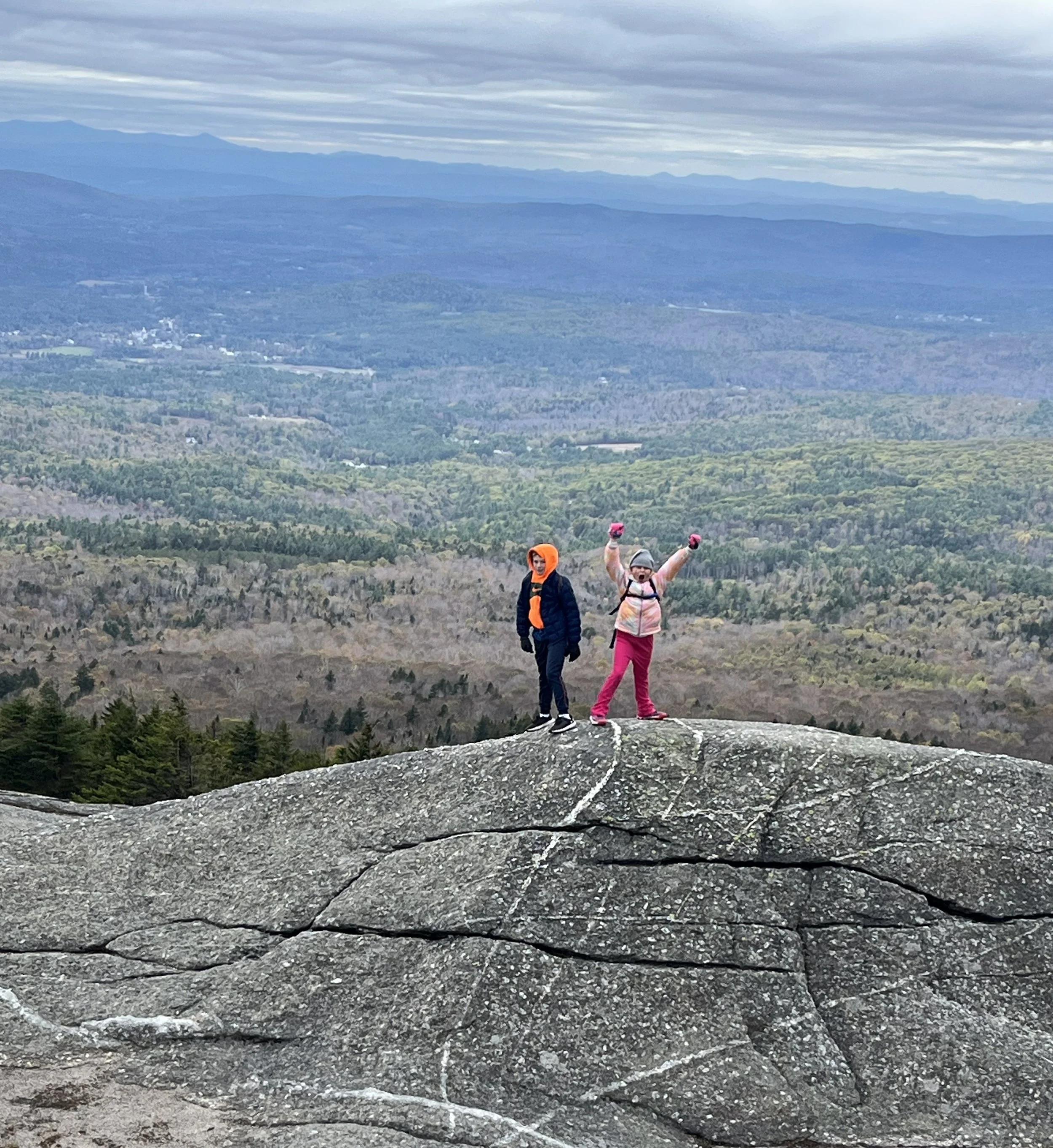 Two children in outdoor clothing and hats standing on a large rock, with a vast mountain landscape and cloudy sky in the background.