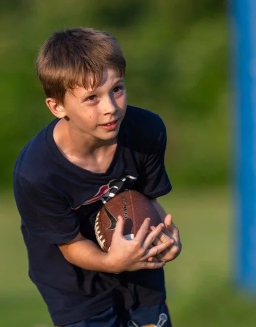 Young boy in a navy T-shirt holding a football, outdoors on a grassy field.