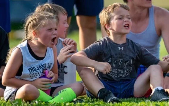Kids sitting on grass, crying, during a sports event, with a woman in the background.