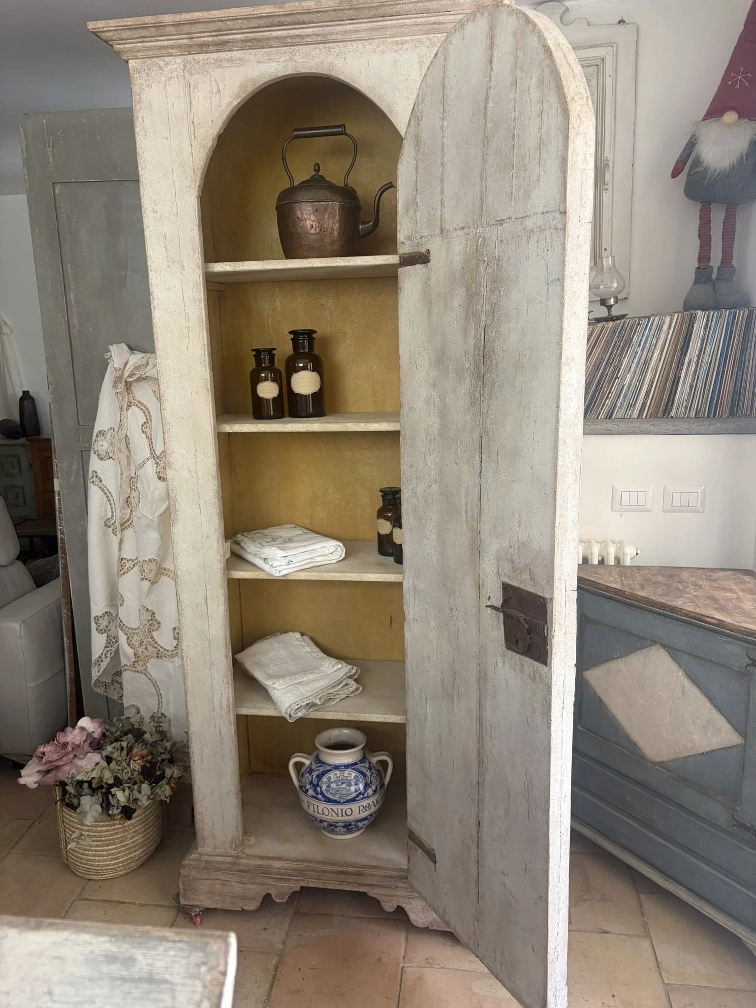 Una vecchia armadio in legno bianco, aperto, con scaffali che contengono oggetti decorativi, tessuti e oggetti d'uso domestico. Sul ripiano superiore una caffettiera di metallo, e sul fondo un grande vaso blu e bianco con scritto 'Filoni Re'. Accanto, si notano altri oggetti in un ambiente domestico.