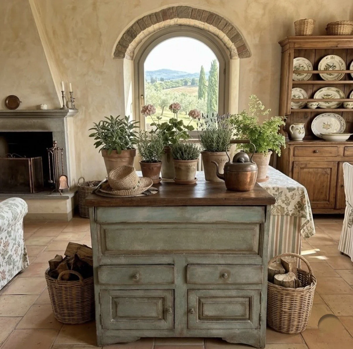 Interno di una cucina rustica con mobili di legno, un camino, piante in terracotta e una finestra che mostra un paesaggio collinare.
