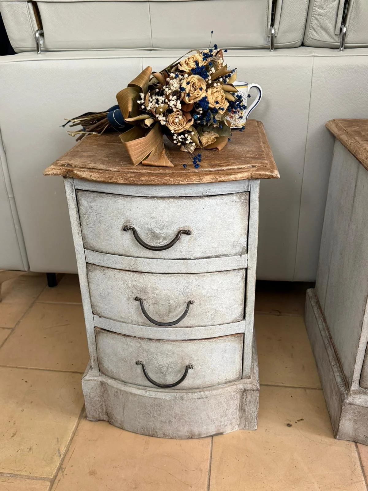Una piccola cassettiera bianca vintage con tre cassetti e maniglie nere, sopra una superficie di legno chiaro. Su di essa è appoggiato un mazzo di fiori secchi, composto da rose beige, piccole fiori bianchi e fiori blu, accompagnato da una tazza bianca con decori blu.