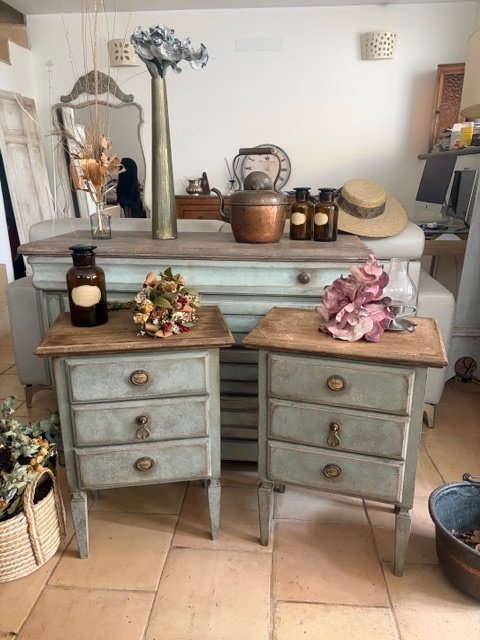 Composizione di arredi e decorazioni vintage con due piccoli comò in stile antico, uno sopra l'altro, decorati con vasi, fiori secchi e oggetti decorativi, ambientata in una stanza con muro bianco e altri mobili in background.