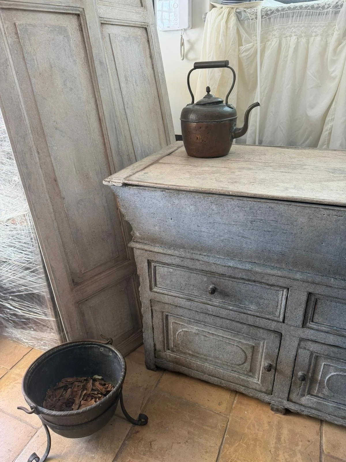 Antico stile mobile in legno con piano in direzione di una parete, con una teiera in metallo scuro sopra. Sul pavimento, un secchio pieno di foglie secche e un sedile con un supporto in metallo.