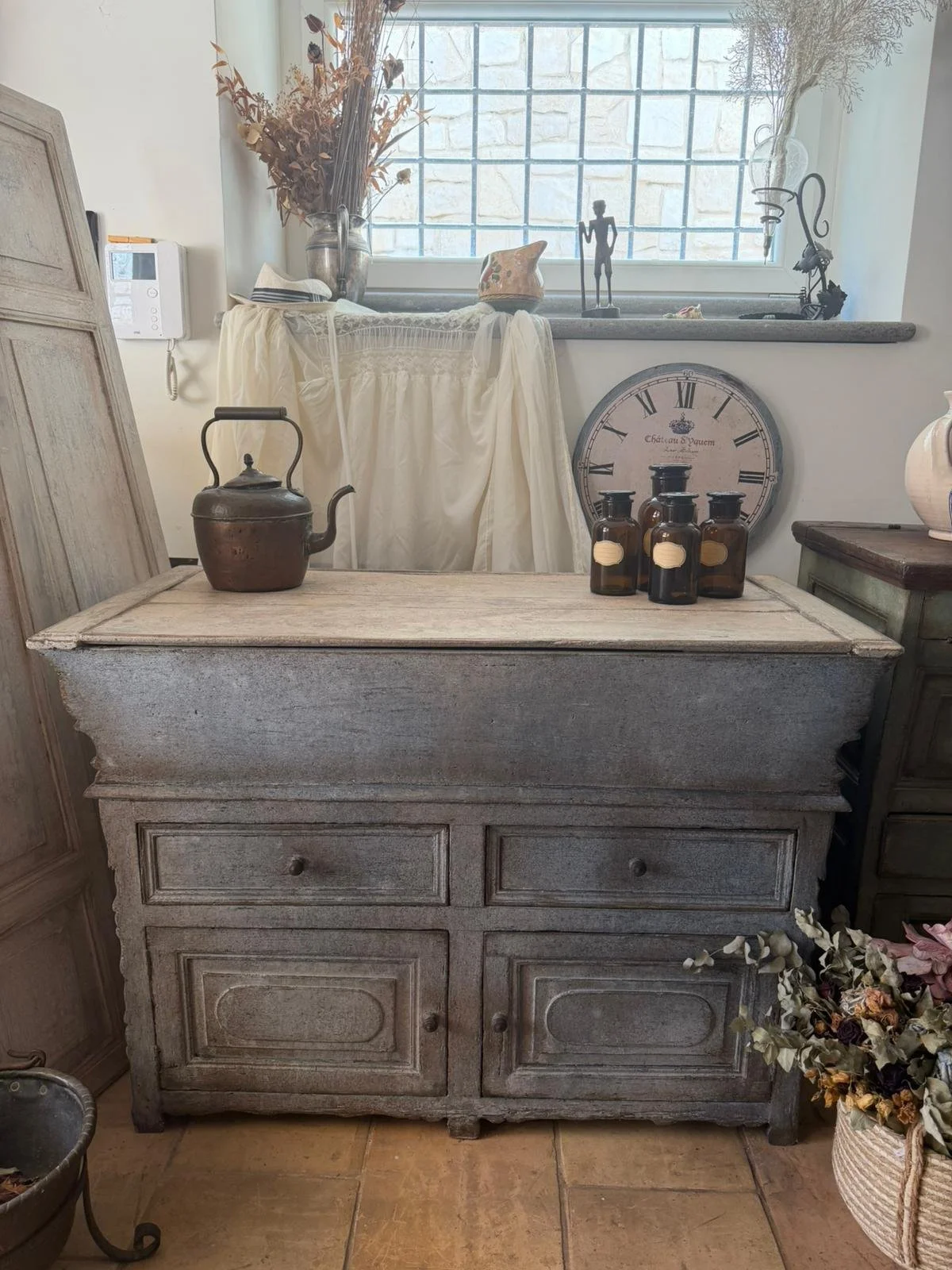 Una credenza in stile vintage con pietra e molti oggetti decorativi, tra cui un tegame di rame, quattro vasi di vetro scuri con etichette, un orologio antico appoggiato sulla parete, e decorazioni come vasi con fiori secchi e statuette, in una stanza luminosa con finestra grande.