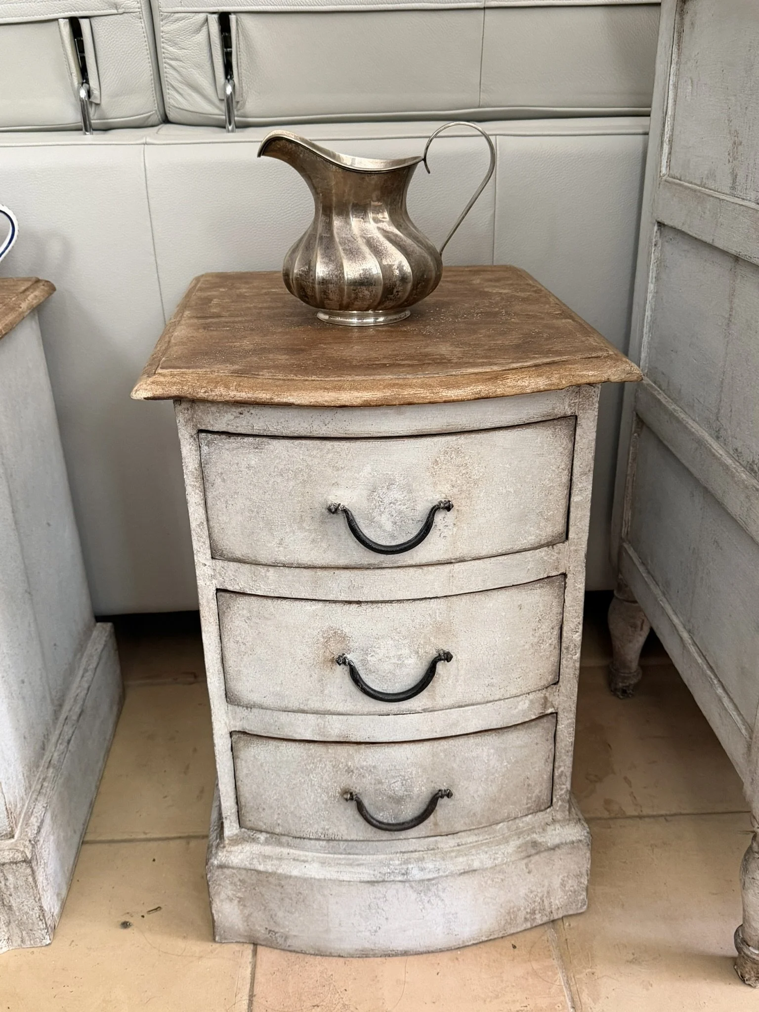 Comò vintage bianco con tre cassetti, in pessimo stato, con maniglie in ferro, in una stanza con pavimento in piastrelle e al lato c'è un mobile parzialmente visibile.