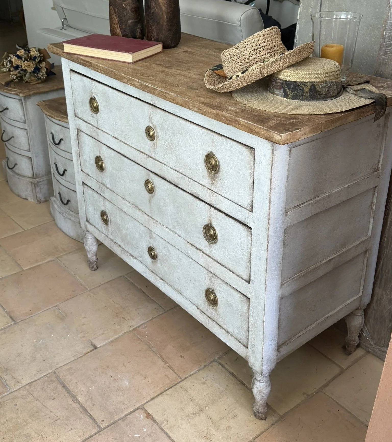 Comò bianco con cassetti, superficie in legno e maniglie in ottone, con cappelli e oggetti decorativi sopra, su pavimento in piastrelle