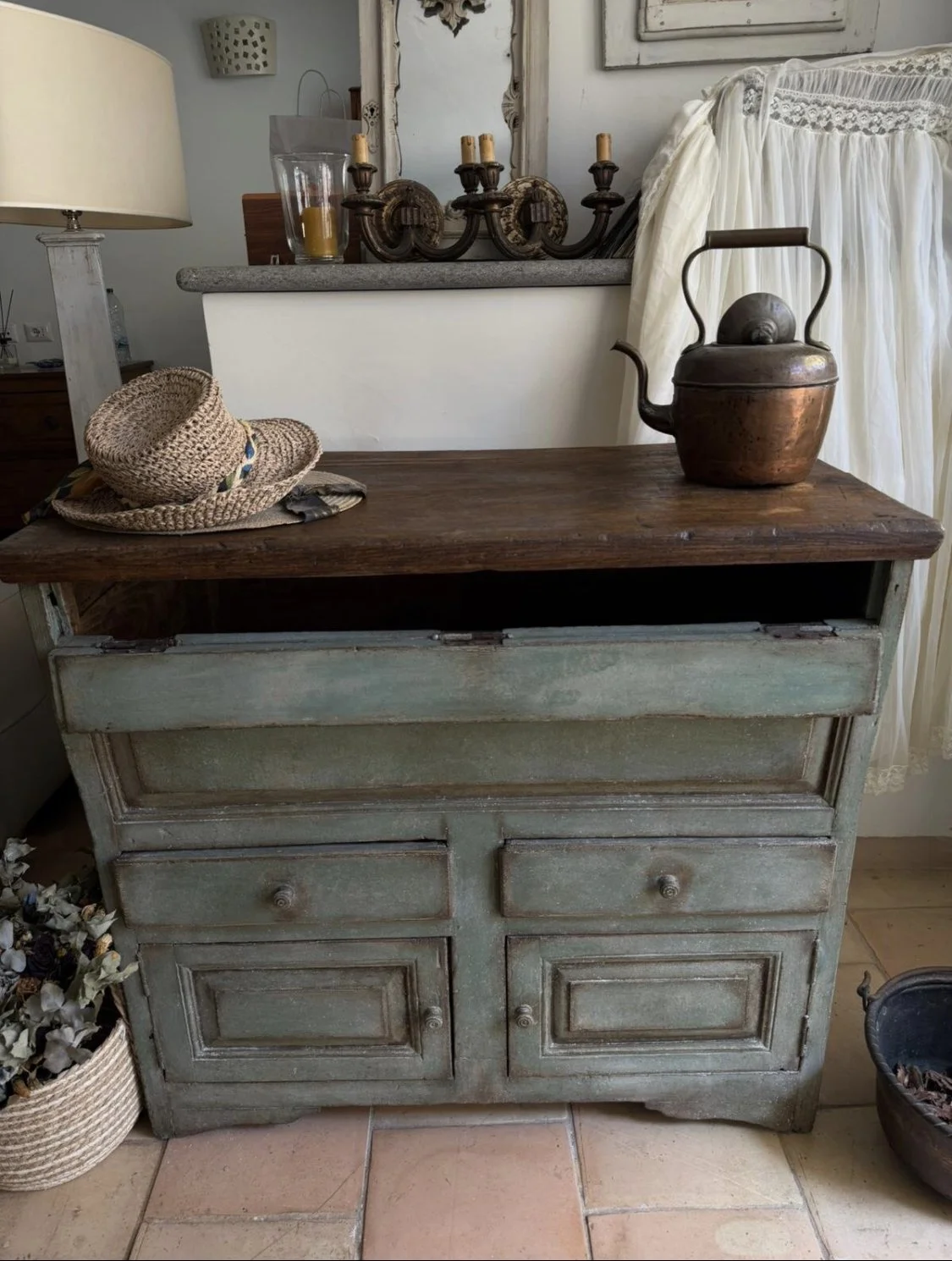 Una credenza vintage con un piano in legno scuro, sopra un cappello di paglia e una teiera in rame. Sullo sfondo, un muro bianco, uno specchio antico, candelabro e lampada con paralume.