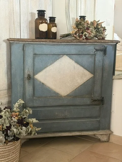 Credenza in legno di colore azzurro con decorazione a rombo sulla porta, decorata con vasi di vetro e fiori secchi sopra, e piante con fiori secchi in un cestino in basso a sinistra.