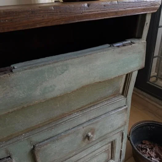 Angolo di un mobile in legno antico con vernice sbeccata, con un cassetto e una credenza inferiore, vicino a una finestra con serranda.