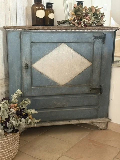 Credenza azzurra con un cassetto decorativo, decorata con bottiglie di vetro marroni e un mazzo di fiori sopra, e un vaso di fiori a terra accanto.