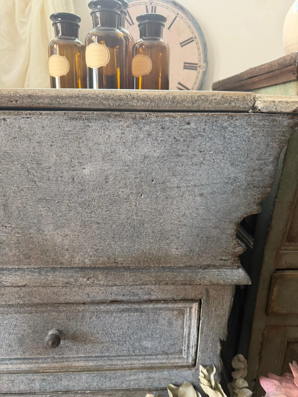 Antico mobile in legno con una finitura invecchiata, con un cassetto e un pomello, sul lato destro si intravede un'altra mobile in legno e sullo sfondo un grande orologio.