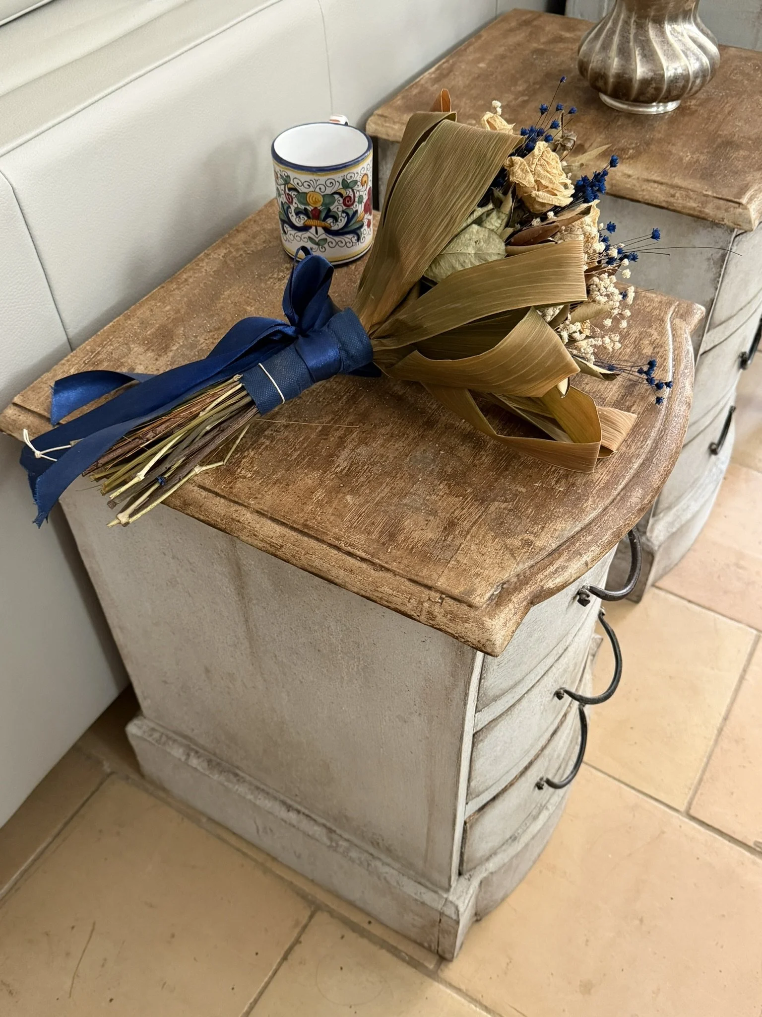 Una composizione di oggetti su un mobile antico in legno chiaro, tra cui un mazzo di fiori secchi e una tazza decorativa con disegni colorati.