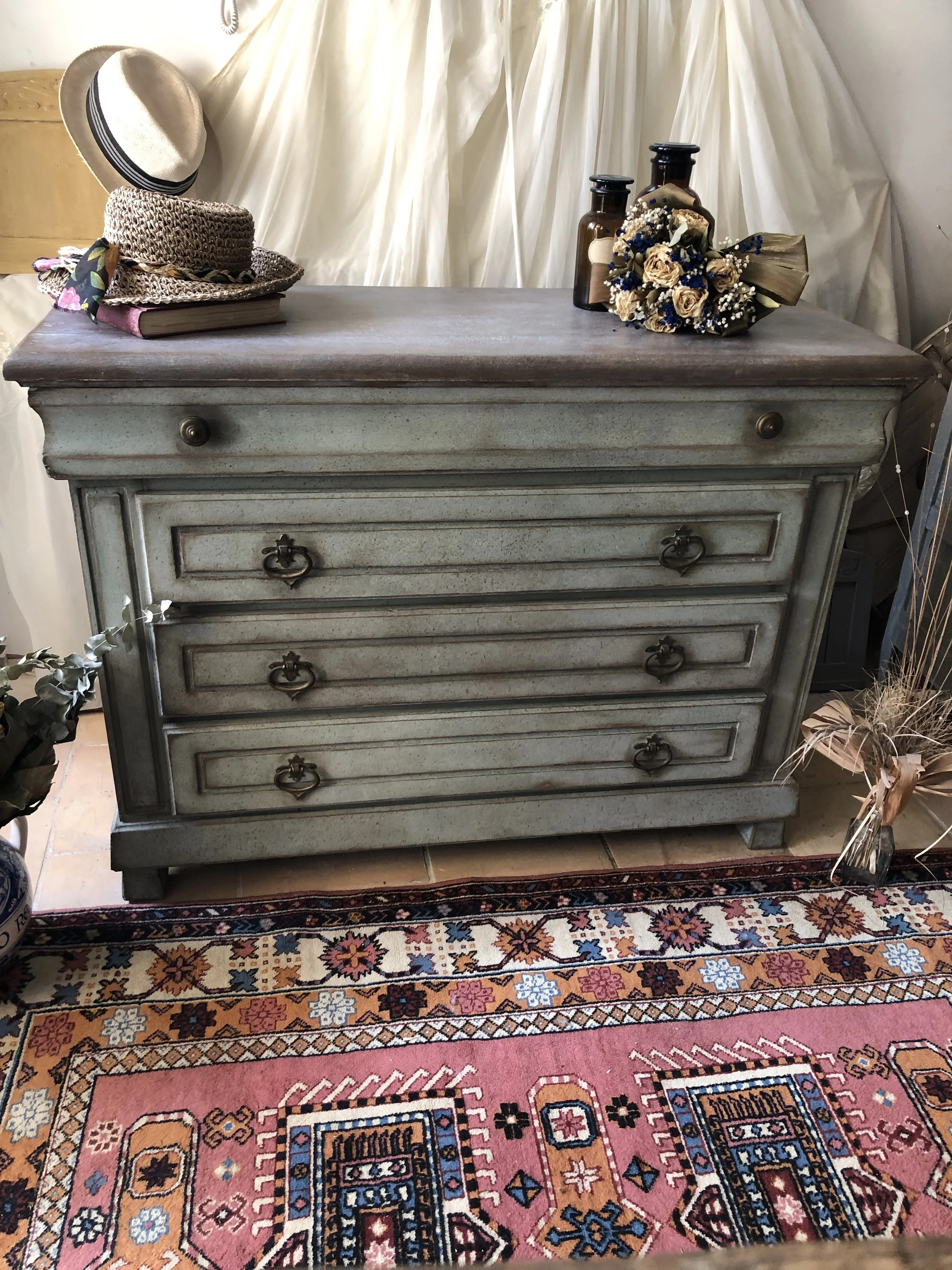 Comò vintage grigio con maniglie in ferro battuto, sotto un tappeto orientale decorato con motivi floreali e geometrici, nello sfondo una tenda beige e oggetti vari.