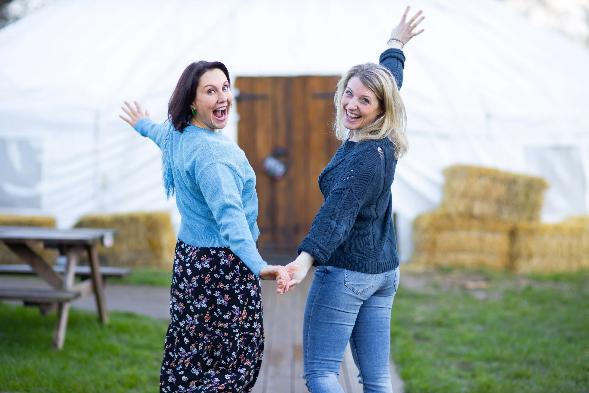 Two women standing outdoors in front of a barn, holding hands, smiling and looking at the camera.