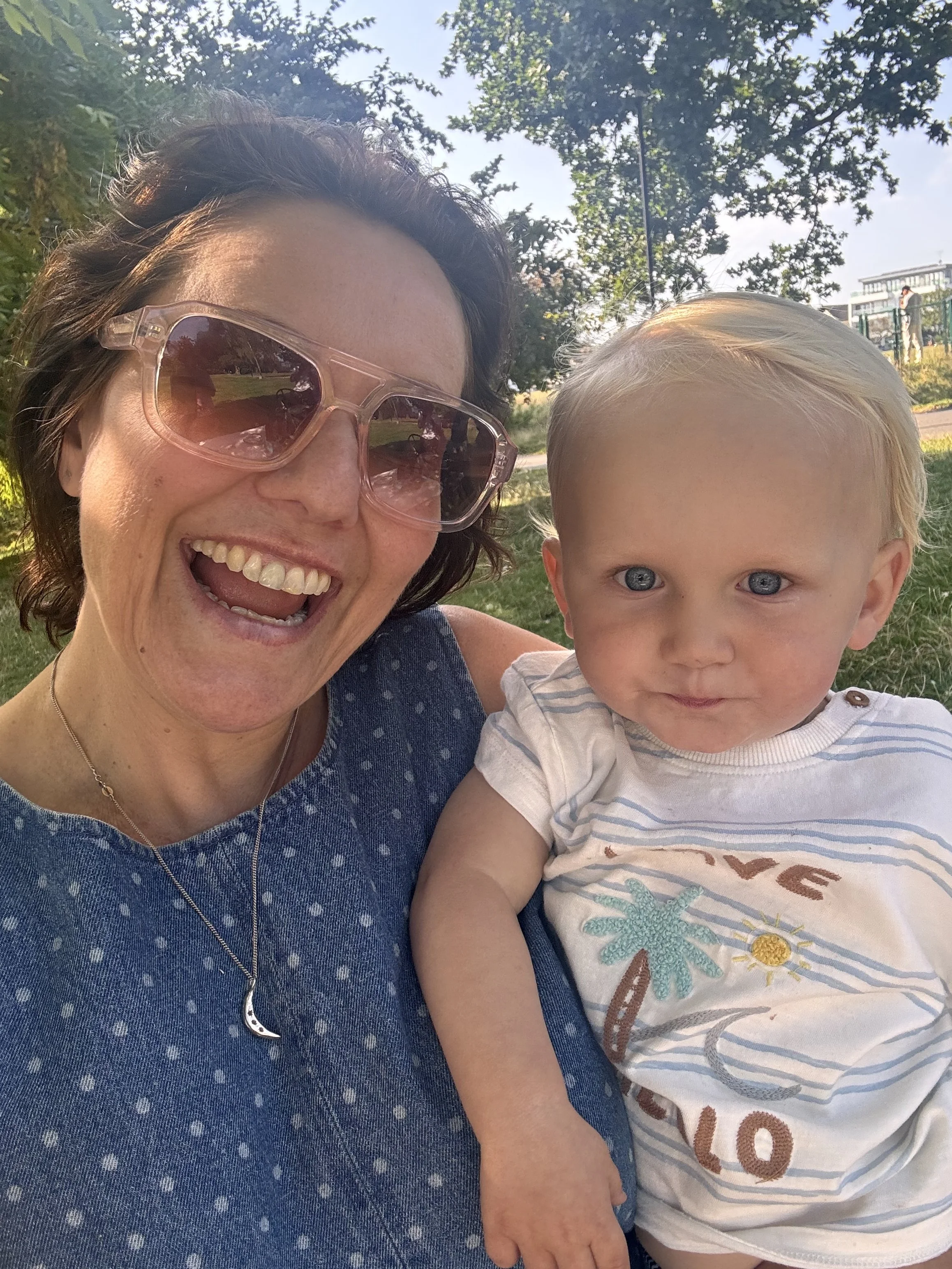A woman with short, dark hair wearing large pink sunglasses, smiling happily, and a young blonde-haired boy with blue eyes, wearing a white t-shirt with colorful graphics, outdoors in a park with trees and a slide in the background.