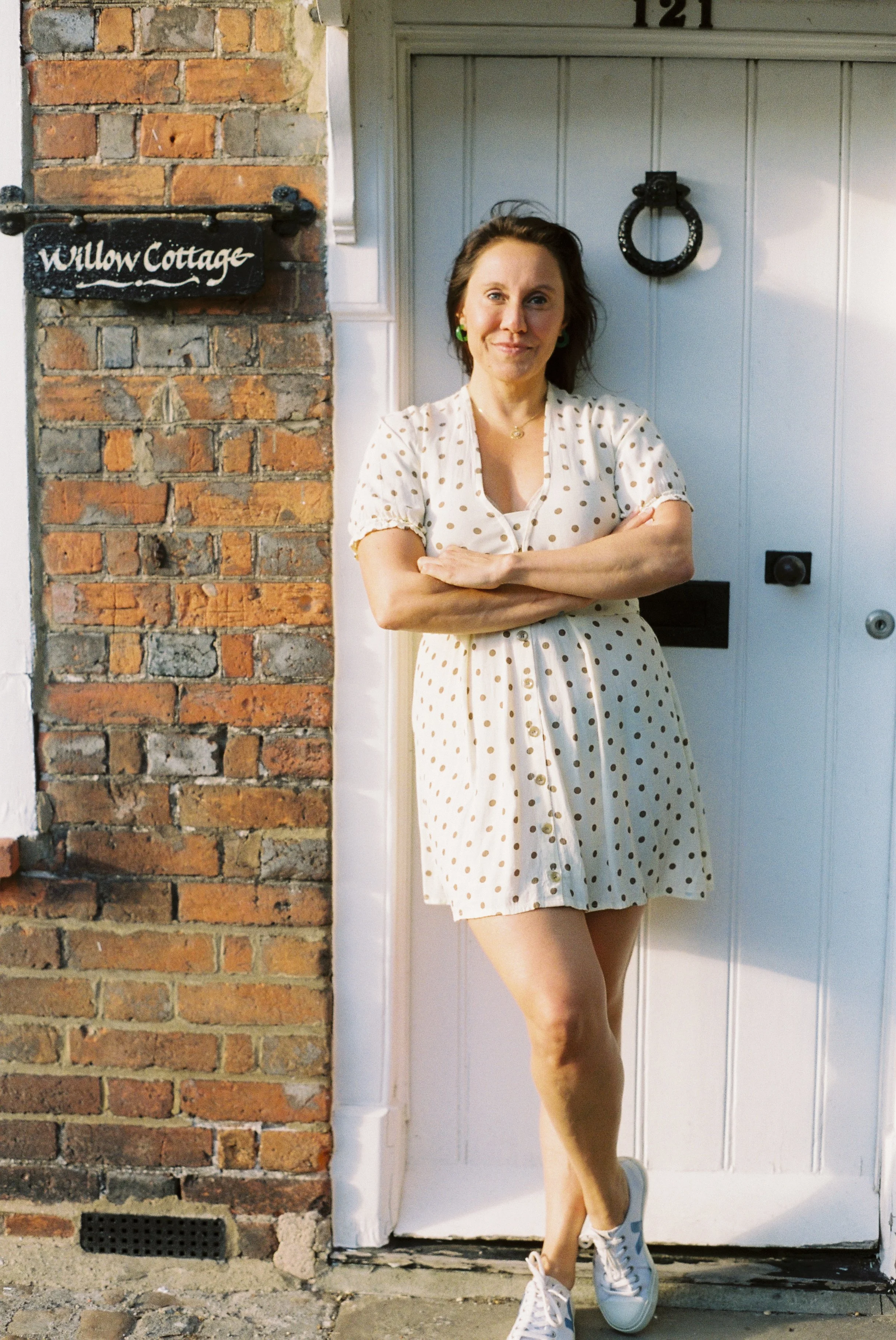 A woman standing with arms crossed in front of a white door, wearing a polka dot dress, white sneakers, and green earrings, beside a brick wall with a black sign that reads 'Wallow Cottage' at the top.