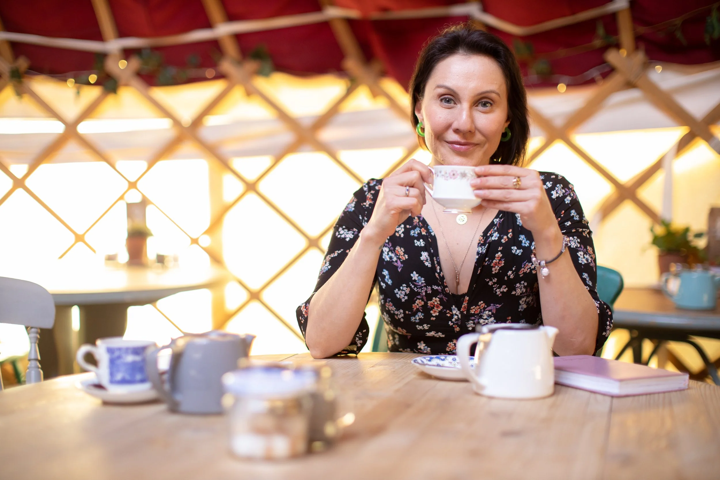 A woman with dark hair and green earrings drinking from a teacup at a wooden table in a decorated, sunlit room.