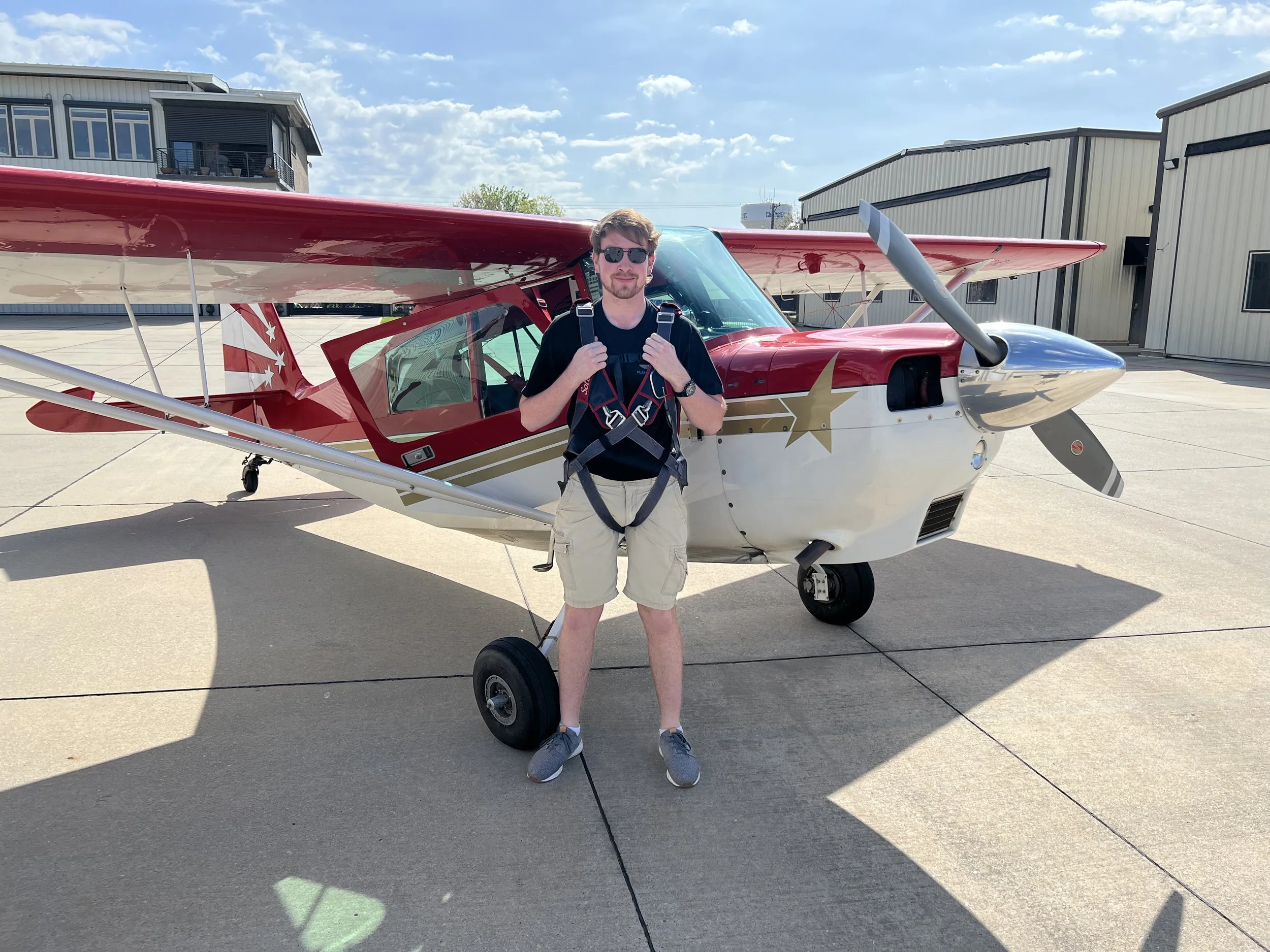 A man with sunglasses, a black t-shirt, and beige shorts standing in front of a small red and white aircraft with a silver nose. He is smiling and holding the straps of his backpack. The airplane is parked on a concrete tarmac, with hangars and buildings in the background under a partly cloudy sky.