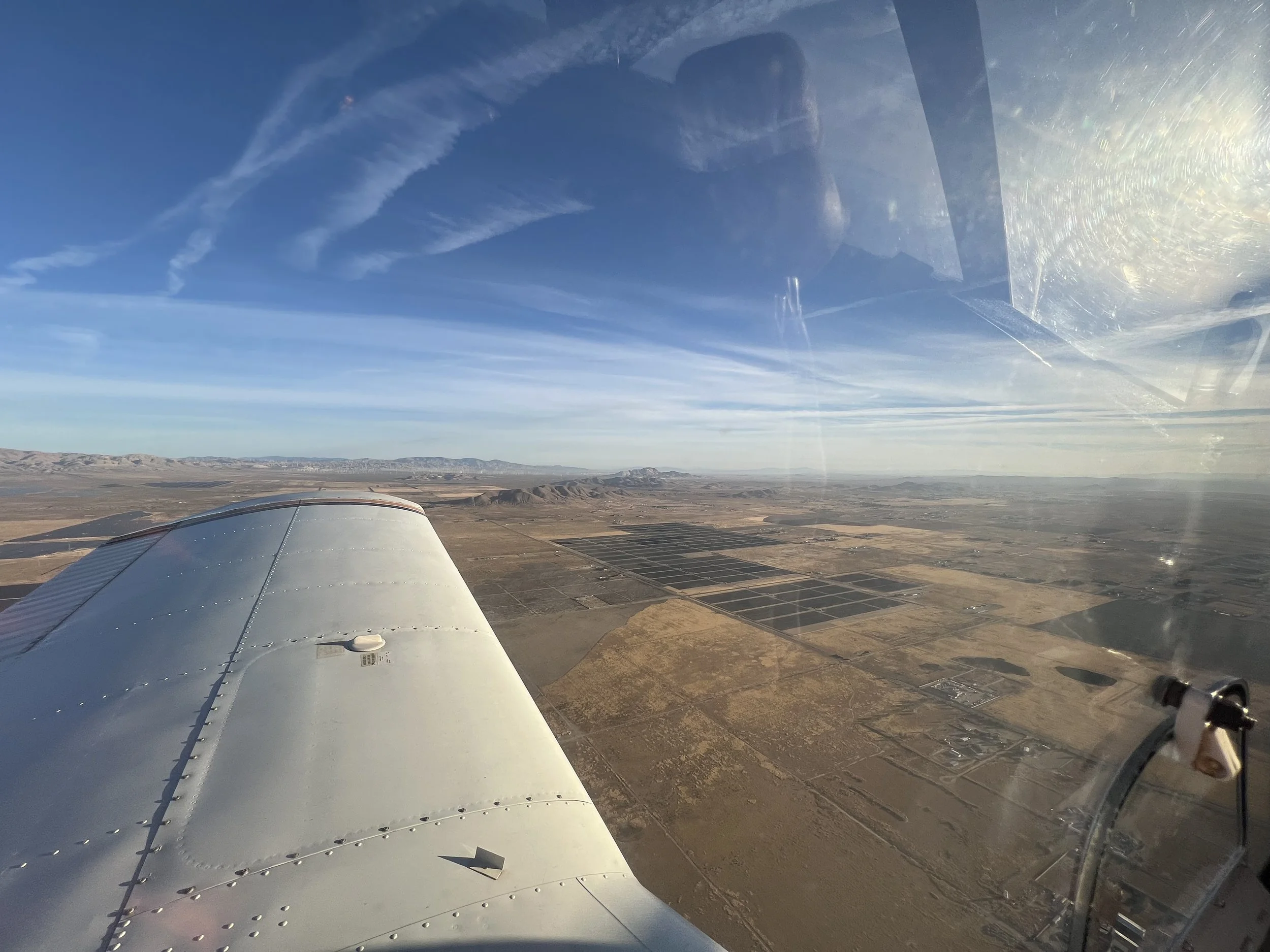 A view from an airplane window showing the aircraft wing over a desert landscape with mountains in the distance under a blue sky with clouds.