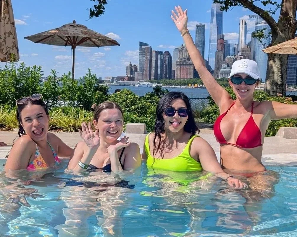 Four women enjoying a pool with a city skyline in the background. They are smiling and making playful faces.