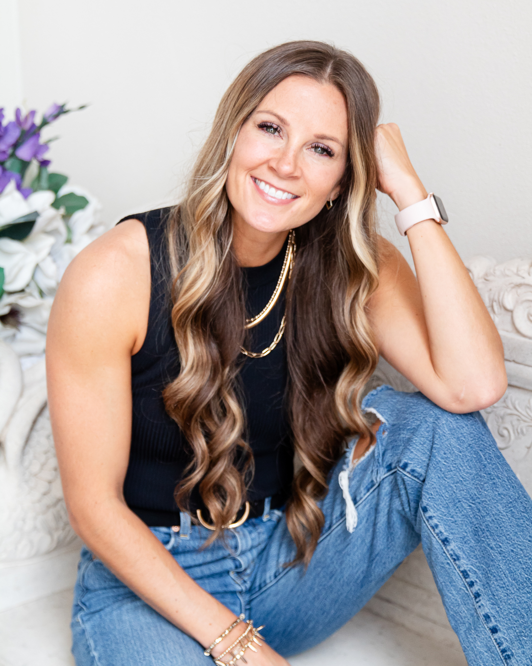 Smiling woman with long wavy brown hair, wearing a black sleeveless top, blue jeans, gold jewelry, and a smartwatch, sitting on the floor indoors next to a white ornate piece of furniture and a bouquet of purple and white flowers.