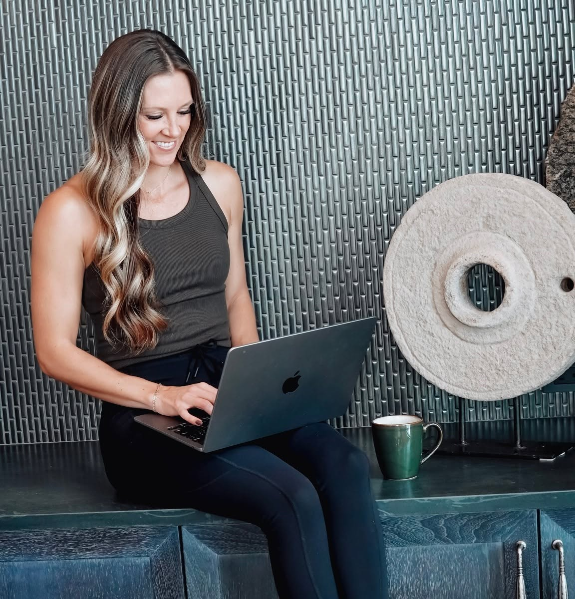 A woman with long wavy hair sitting on a dark table, smiling while using a laptop. She is wearing a sleeveless black top and black pants. Next to her on the table is a green mug. Behind her is a textured wall with decorative objects, including a large stone or ceramic disc and a white stone sculpture.