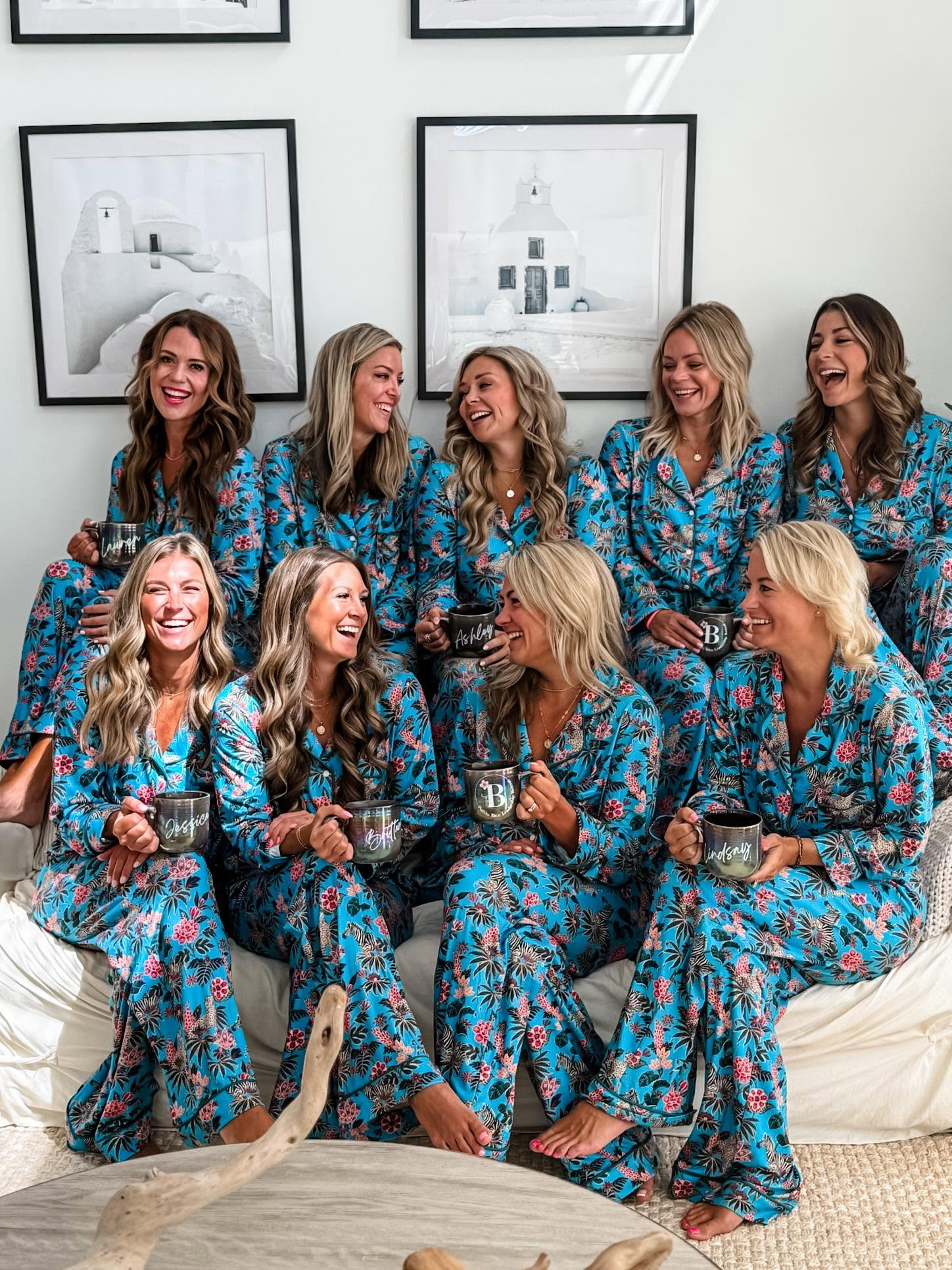 Group of women in matching blue floral pajamas sitting on a couch, laughing and holding personalized coffee mugs. Some women are seated, some are standing behind, with a wall adorned with black and white framed artwork of white buildings in the background.