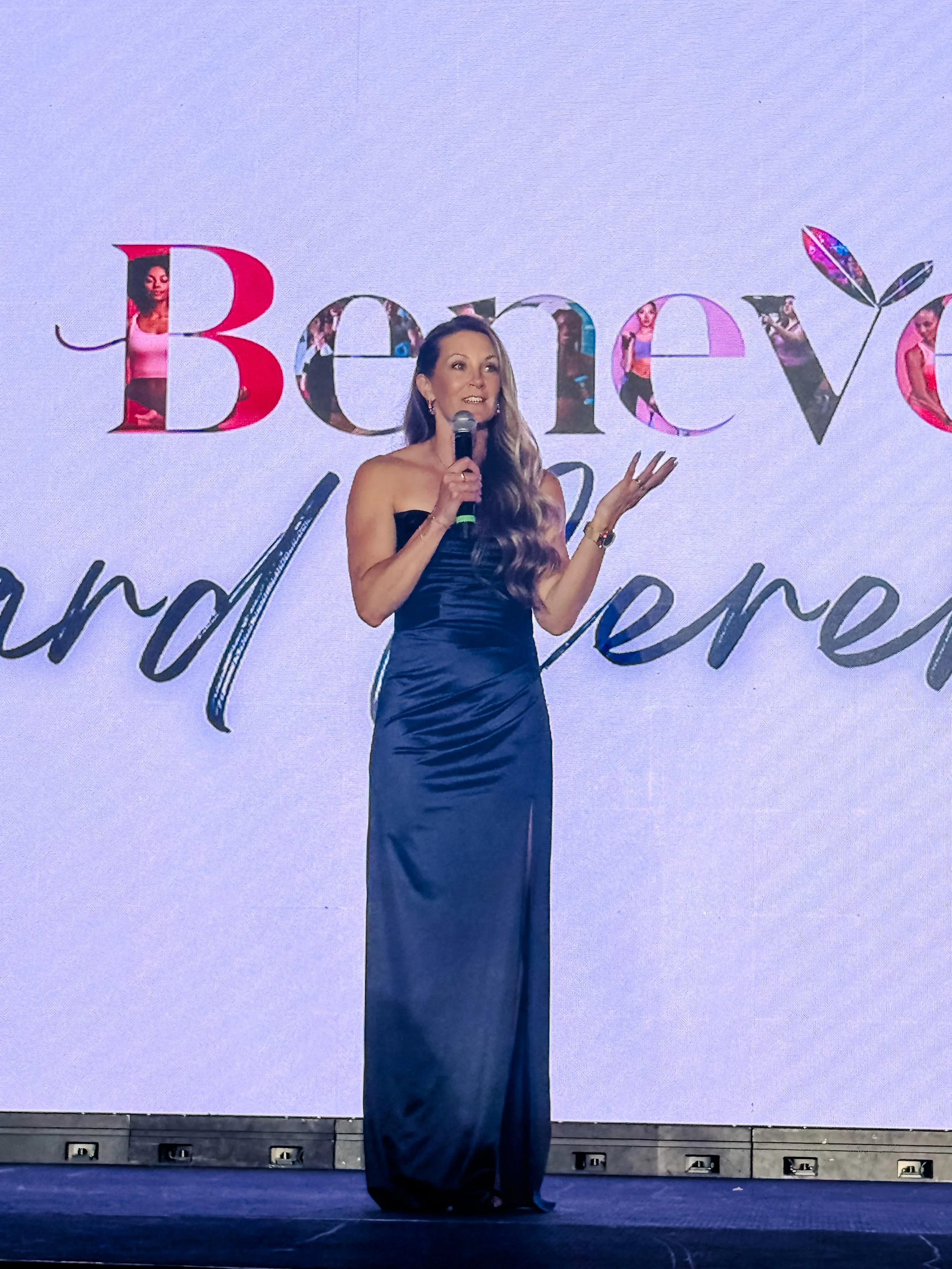 A woman in a long black dress speaking into a microphone on stage during a formal event with a large screen behind her displaying the partially visible phrase "Be..." in stylized text.